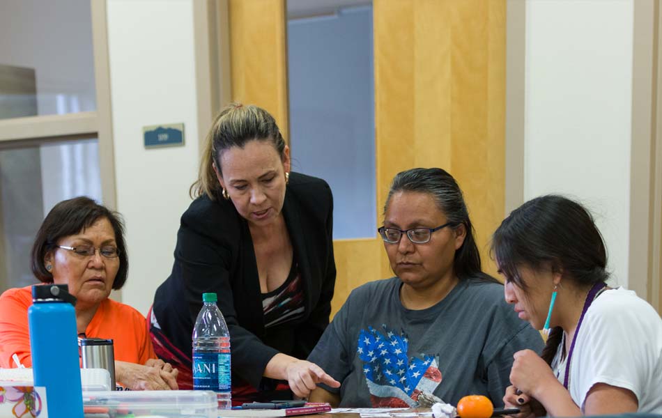 Facilitator leaning over a table to guide a small group of adults reviewing printed materials together in a classroom or workshop setting.