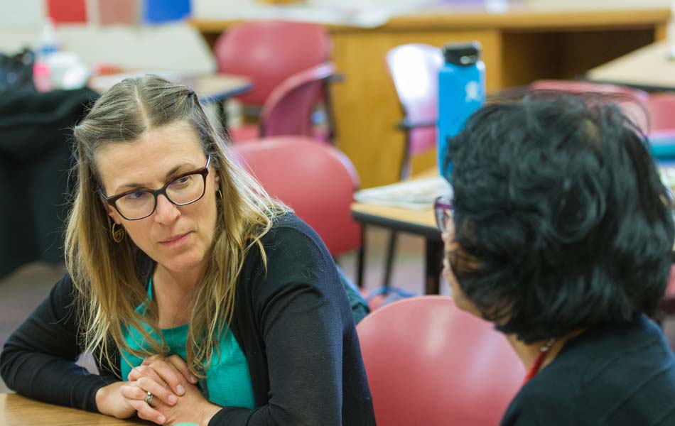 Two adults seated at a classroom table engaged in focused conversation, with notebooks, chairs, and classroom furnishings visible in the background.