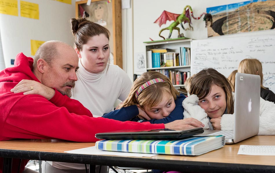 Group of adults and children gathered around a table in a classroom, working together on a laptop with books, notebooks, and classroom materials visible.