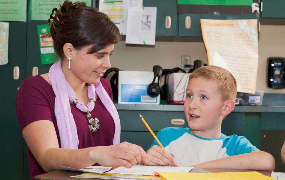 Adult working one‑on‑one with a child at a classroom table, reviewing worksheets and writing with a pencil, with classroom materials and posters in the background.