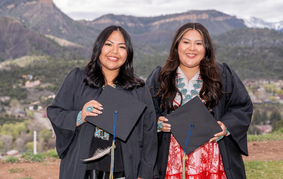 Two graduates wearing caps and gowns, holding diplomas, standing outdoors with mountain scenery in the background.